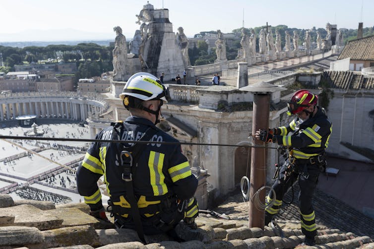 Dois bombeiros em um telhado com vista para a Praça de São Pedro.